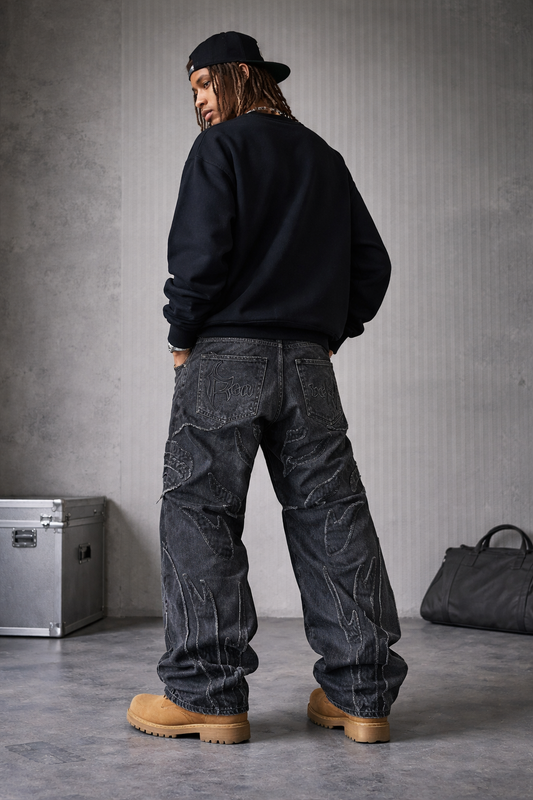 Person wearing a black sweatshirt and dark jeans standing against a gray wall with a suitcase and bag on the floor.